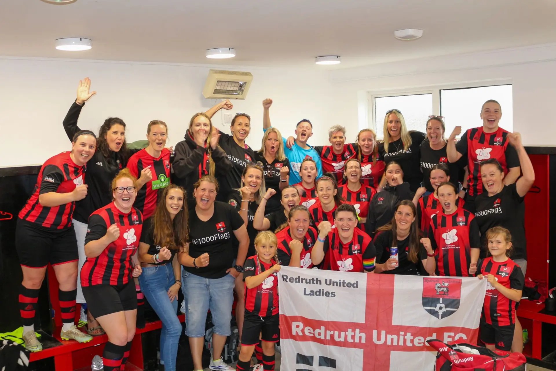 Redruth United Ladies celebrating together in the changing room with a club flag