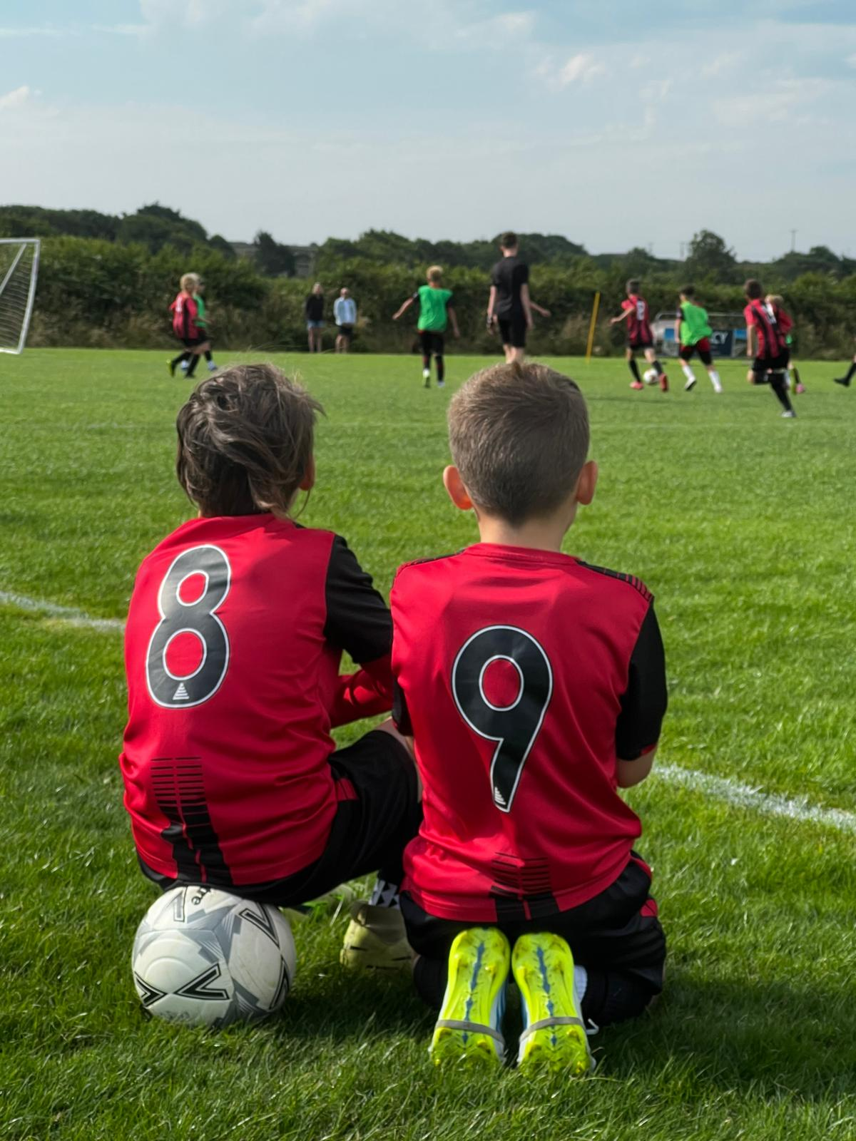 Two young Redruth United players in red and black kit watching a match from the sideline